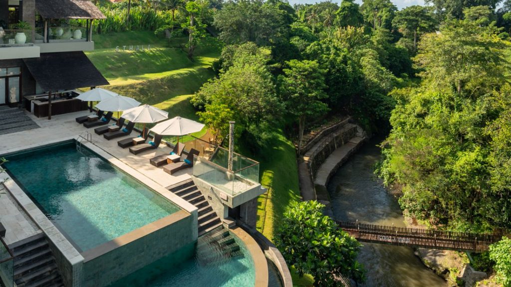 Luxury infinity swimming pool overlooking a lush tropical river valley, with sun loungers and umbrellas at a Bali resort.
