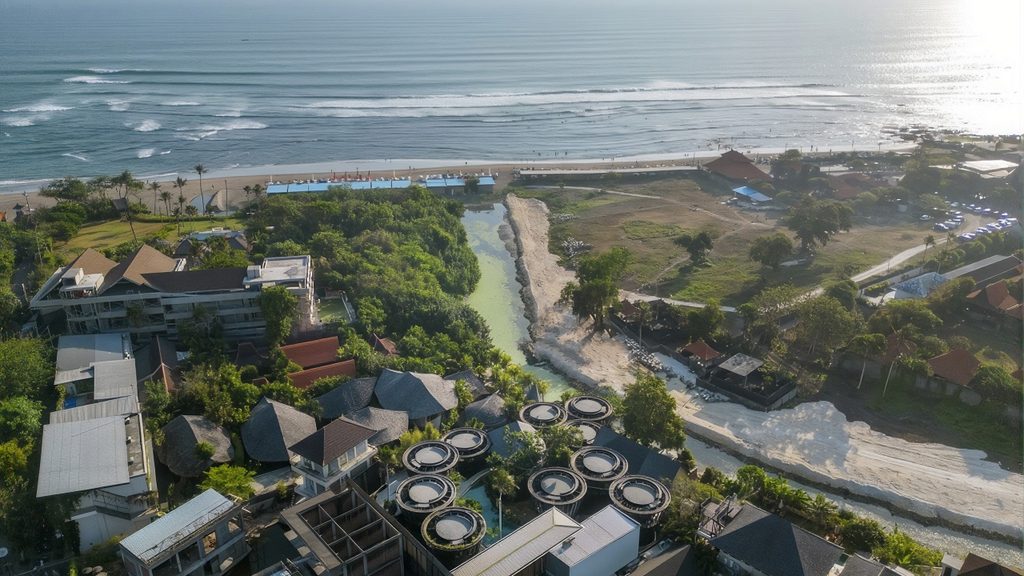 Aerial view of a beachfront resort area in Bali, featuring tropical buildings, a winding river, and ocean waves along the sandy coast.