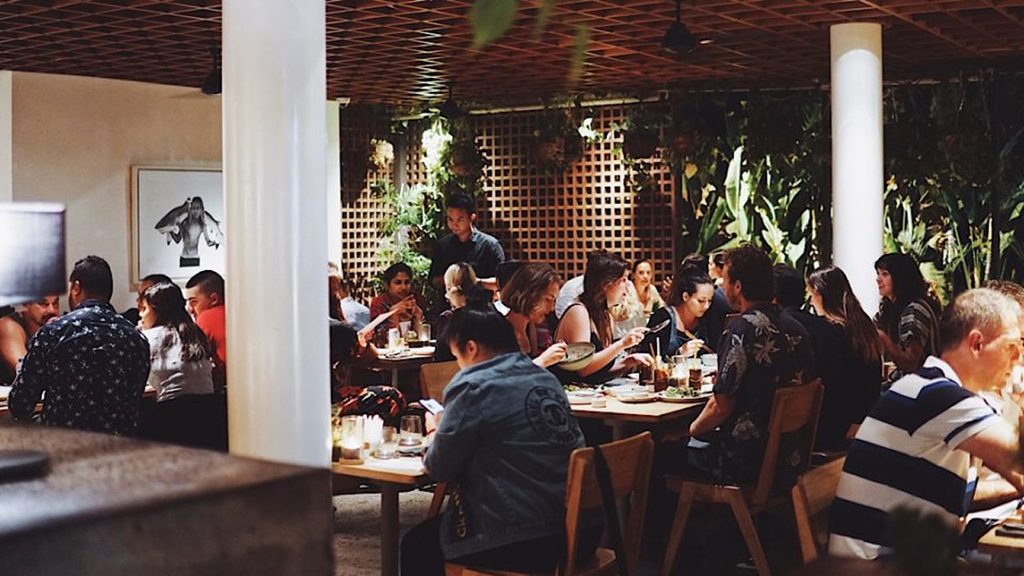 Busy restaurant interior with guests enjoying food and drinks at wooden tables, surrounded by tropical greenery and warm lighting.