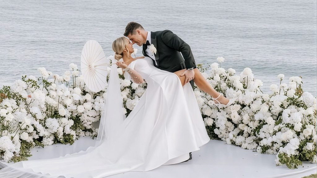 Bride and groom sharing a romantic moment surrounded by elegant white floral arrangements with an ocean backdrop.