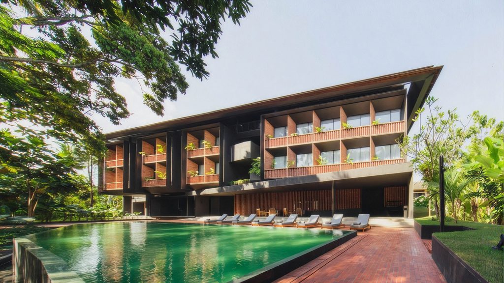 Modern luxury resort building with wooden balconies, a large outdoor swimming pool, and sun loungers surrounded by tropical greenery.