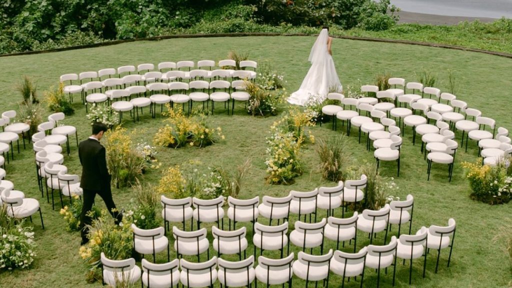 Outdoor garden wedding ceremony with circular seating arrangements, white chairs, yellow floral decorations, and a bride and groom on a green lawn.
