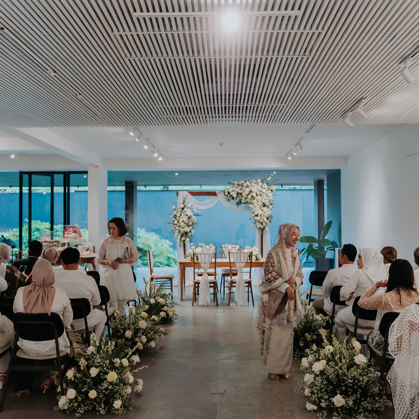 Wide view of a modern indoor wedding ceremony with white floral decor and architectural slatted ceilings at Titik Du